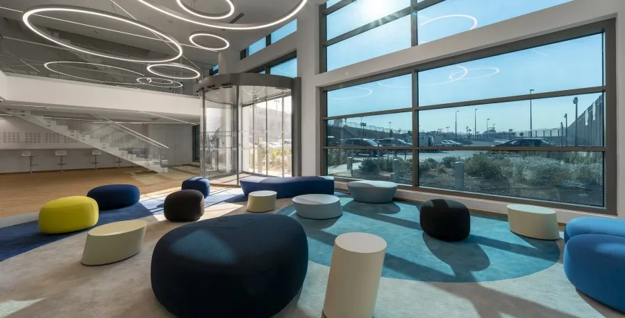 View of a big, brightly lit room, looking towards a whole wall of external windows and a glass door vestibule. Circular artful lighting on the ceiling, a glass stairway on the farthest wall with maybe some stools under it, and then the floor is mostly carpeted with a bunch of cushy looking bean bags and small pedestal tables are placed sporadically around the sunlit room.