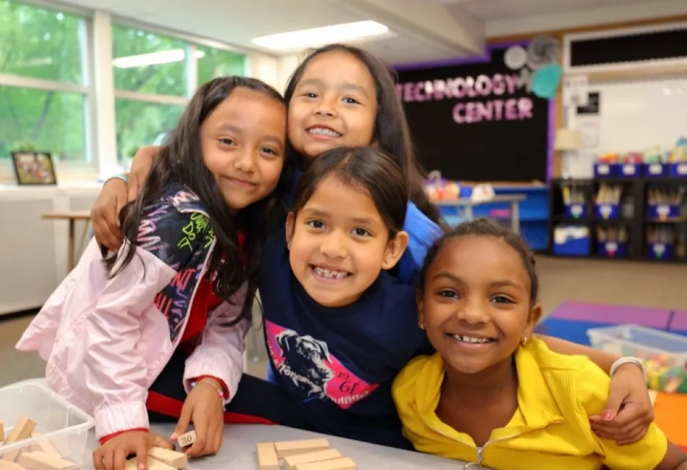 Image of 4 young kids hugging, in a classroom, huddled around a table playing blocks. They look like they're all friends.