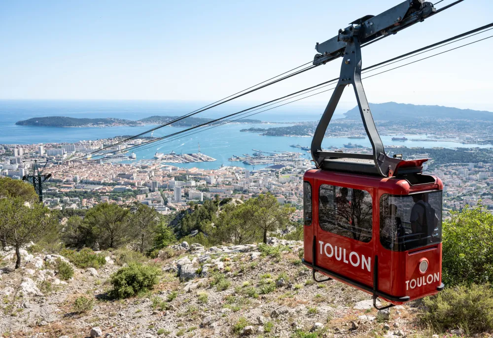A panoramic view of the city of Toulon, featuring a red cable car with "Toulon" written on it in the foreground and the sea in the background.