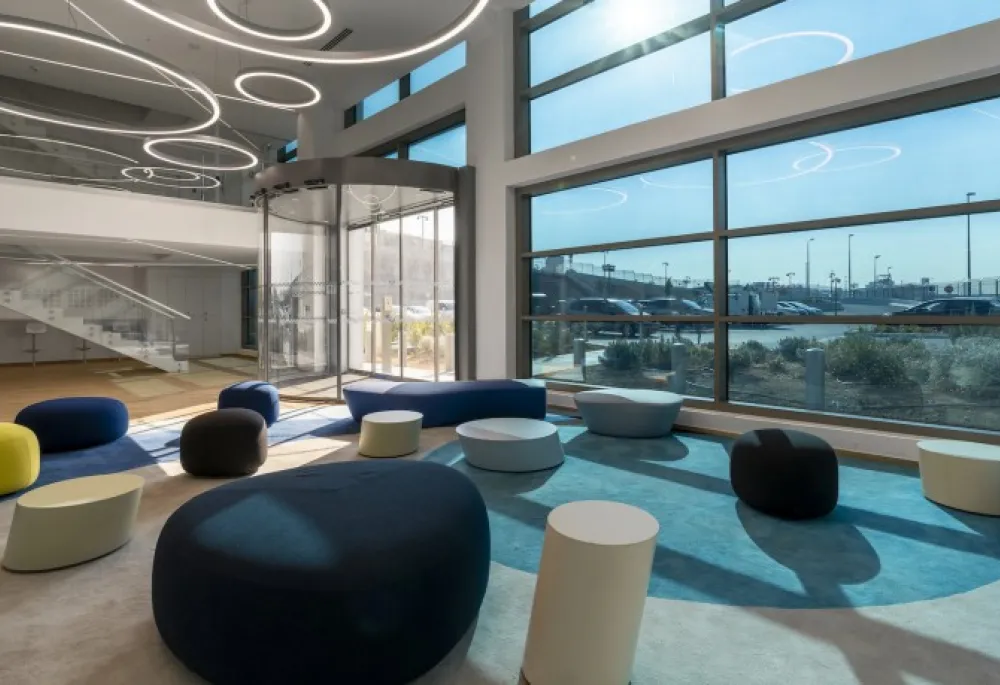 View of a big, brightly lit room, looking towards a whole wall of external windows and a glass door vestibule. Circular artful lighting on the ceiling, a glass stairway on the farthest wall with maybe some stools under it, and then the floor is mostly carpeted with a bunch of cushy looking bean bags and small pedestal tables are placed sporadically around the sunlit room.