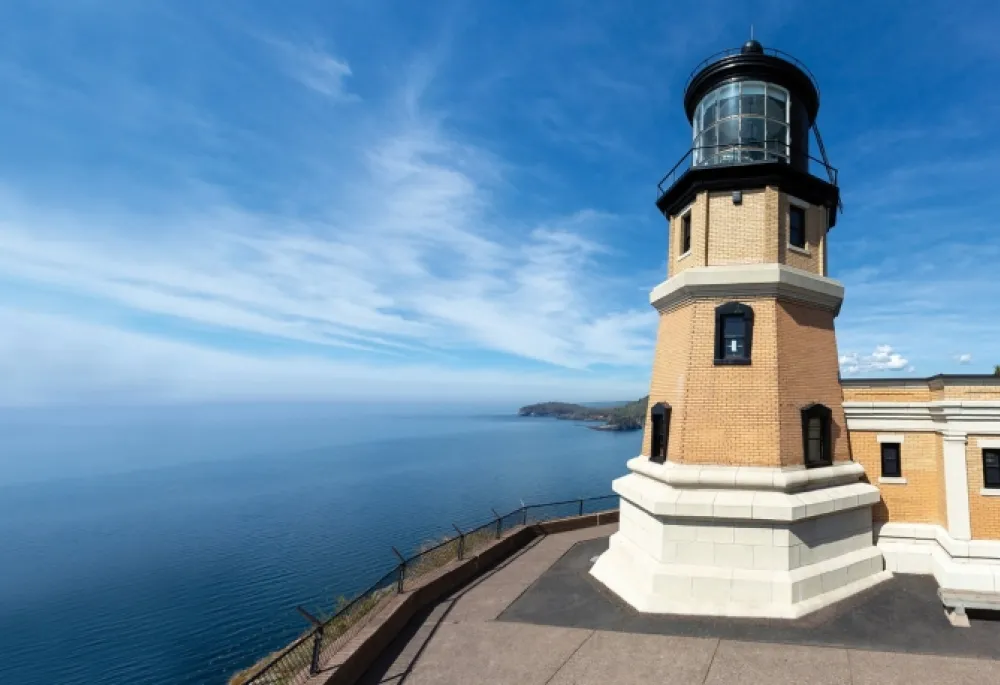 Image of Split Rock Lighthouse, up close. It appears to be up on the top of a hill, overlooking water and the sky, and there's a couple story tall lighthouse that is 20 feet away.