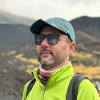 A man in a billed cap in a yellow jacket. There are hills behind him. He wears sunglasses.
