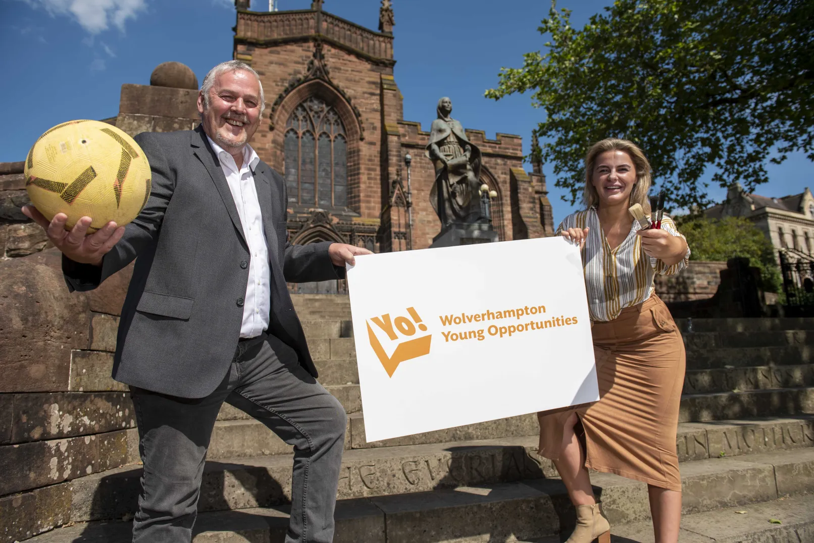 Two councillors outdoors holding a Yo! Wolverhampton Young Opportunities sign, a football and paintbrushes