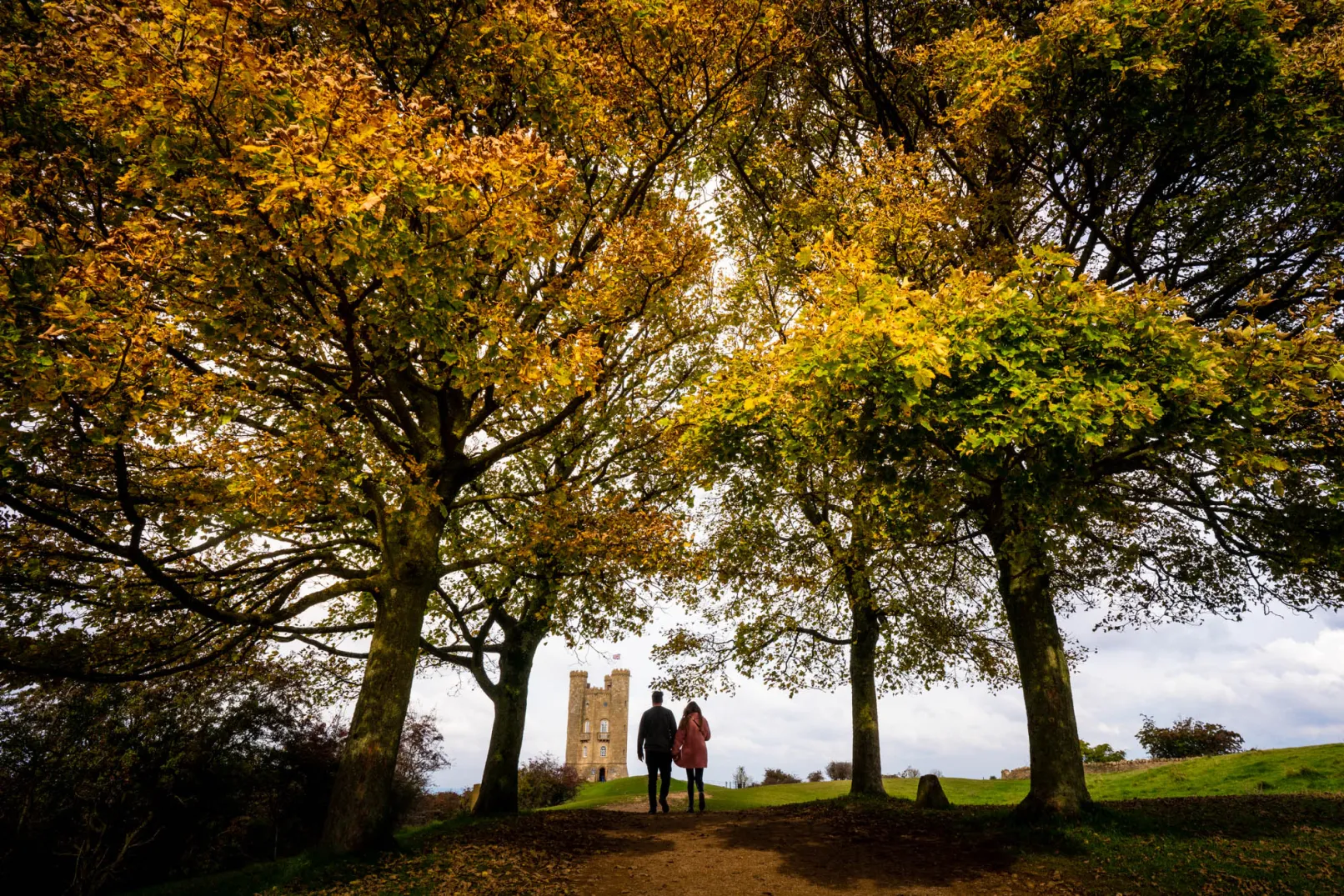 Two people walking beneath autumn trees towards Broadway Tower in Worcestershire