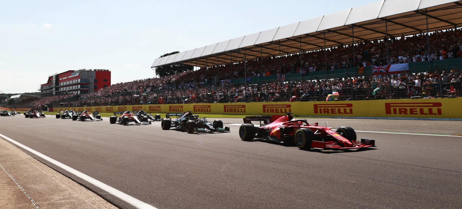 Formula 1 cars parading in formation, starting with the gleaming red Ferrari, in front of the Silverstone circuit's grandstand, which is full of fans.